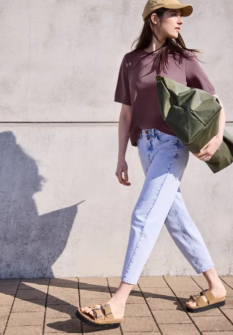Frau in lockerem T-Shirt, hellen Jeans und Sandalen mit großer grüner Tasche vor heller Wand.